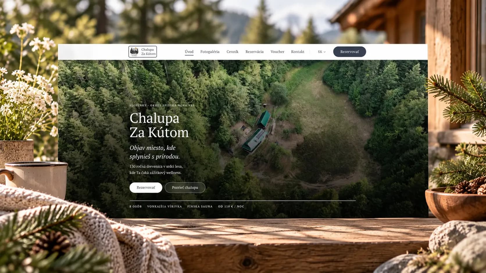 The Chalupa Za Kútom home page with an aerial view of the forest on a screen on a wooden porch table, a mug, a blanket and fir twigs beside it